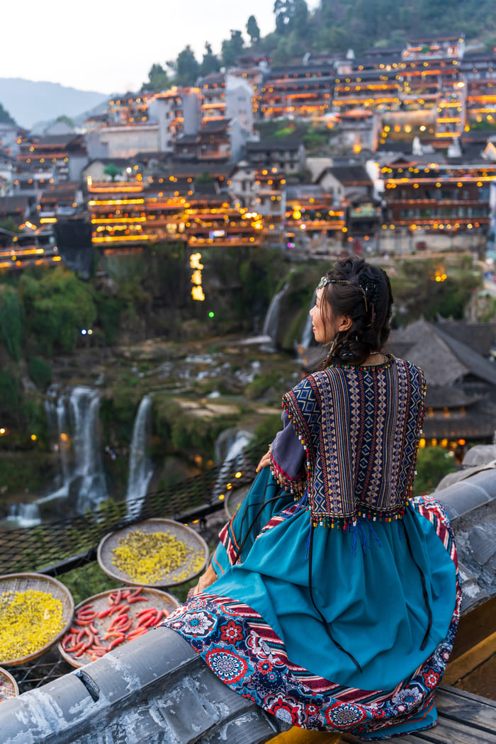 Una chica con roopa tradicional sentada en un peldaño admirando el paisaje, con un pueblo tradicional en las montañas de fondo